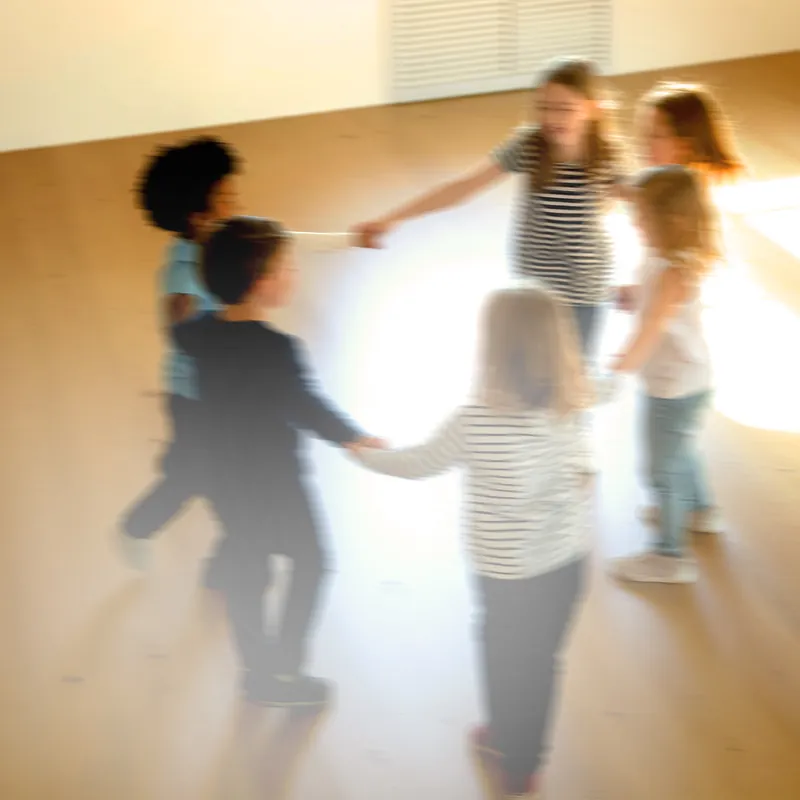 Kinder fassen sich an den Händen und drehen sich gemeinsam im Kreis in einem hellen, sonnigen Raum.